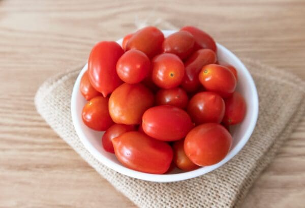 TOMATOES MINI ROMA White bowl with small red tomatoes on burlap and wooden surface.
