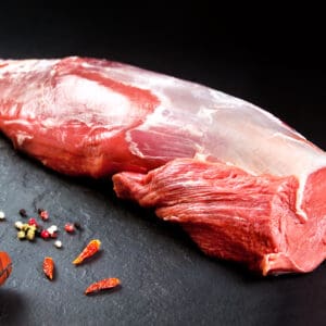 Raw red meat on dark surface with knife, chili peppers, peppercorns scattered nearby.
