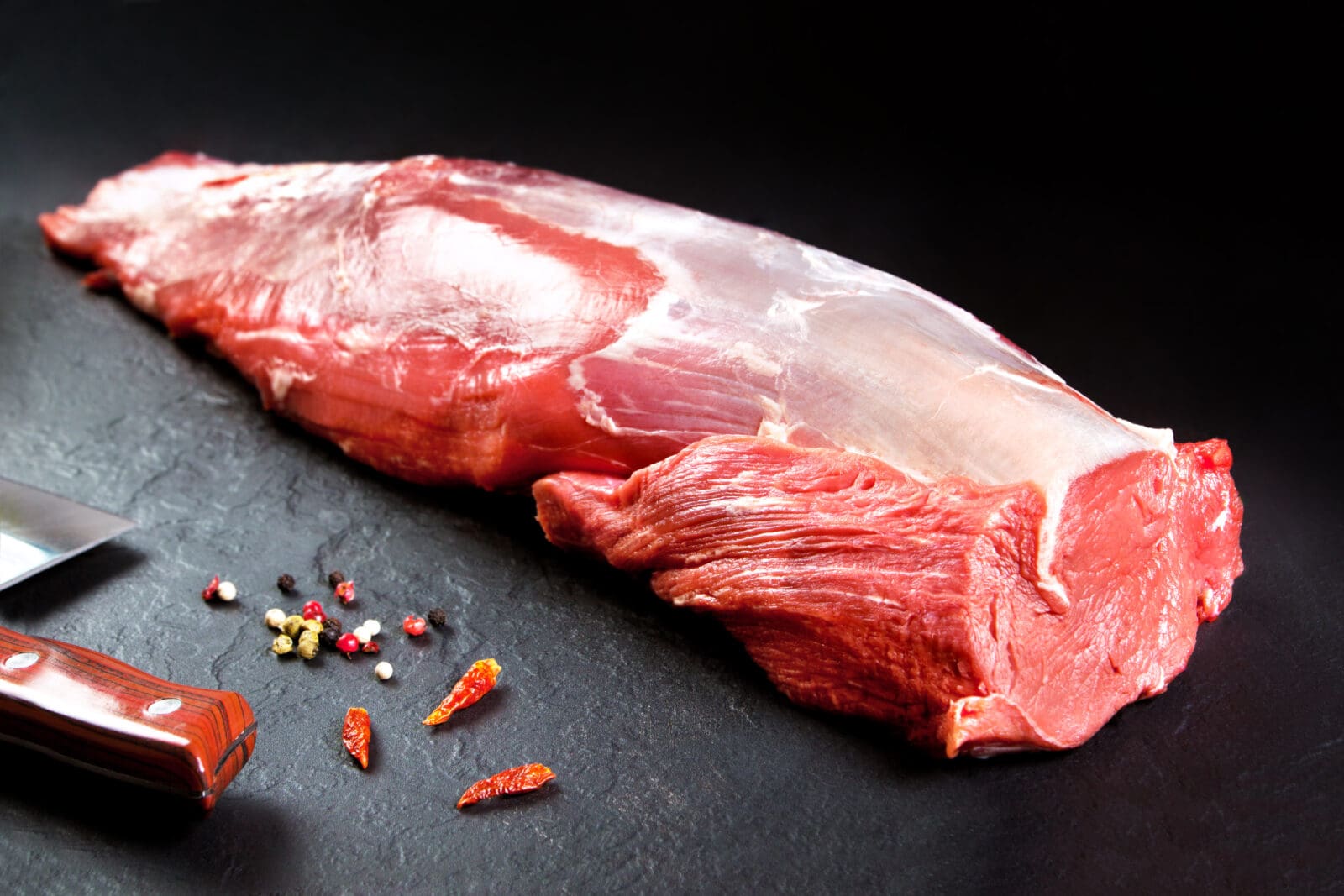 Raw red meat on dark surface with knife, chili peppers, peppercorns scattered nearby.
