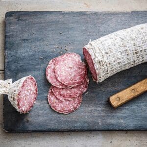 Partially sliced salami wrapped in netting with round slices and knife on a wooden board.