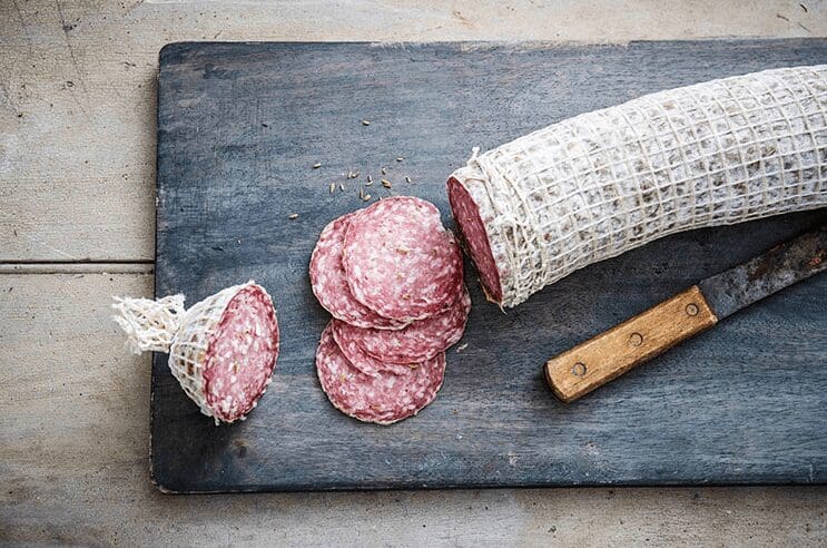 Partially sliced salami wrapped in netting with round slices and knife on a wooden board.