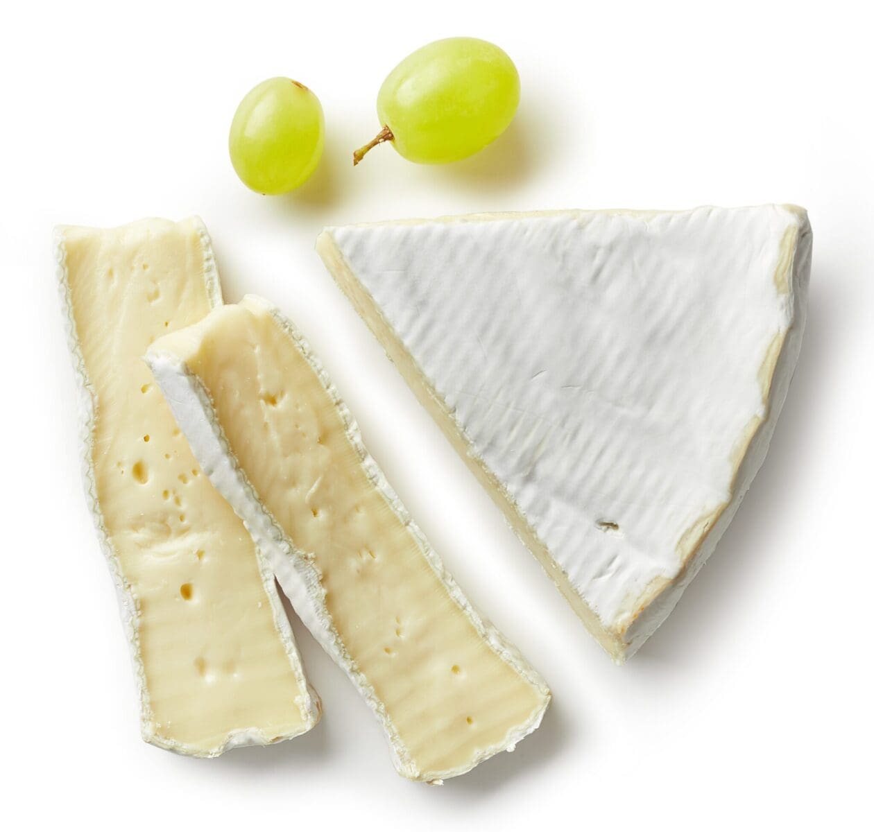 Wedge and slices of soft cheese with rind and two green grapes on white background.