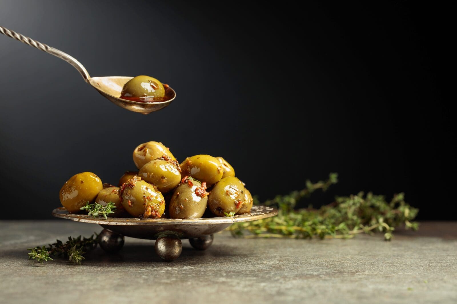 Spoon holds a marinated green olive above a plate of seasoned olives with herbs.