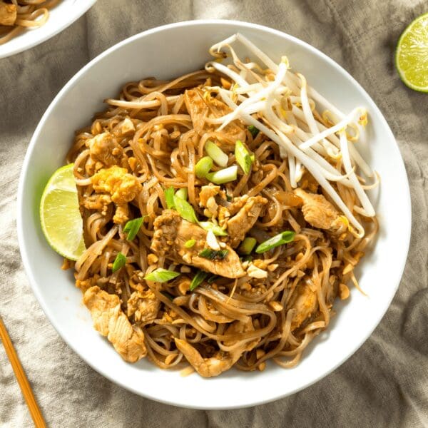 Bowl of stir-fried noodles with chicken, bean sprouts, green onions, peanuts, and lime wedge.